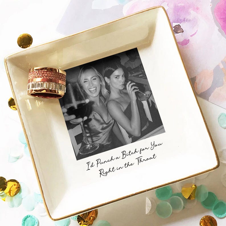 May include: A square, off-white trinket dish with a gold rim, featuring a black and white photo of two women and the text "I'd Punch a Bitch for You Right in the Throat." A rose gold ring sits on the dish.