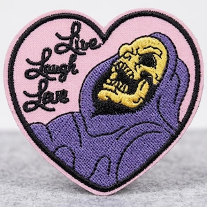 May include: A heart-shaped patch with a pink background and a black border. The patch features a purple hooded figure with a yellow skull and the words "Live Laugh Love" in black script. The patch is likely an iron-on or sew-on.