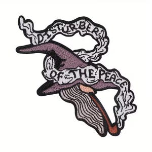 May include: Embroidered patch featuring a witch's hat in shades of purple and black, with a brown stem. The patch includes the text "Disturber of the Peace" in a white, stylized font, surrounded by a white smoke design.
