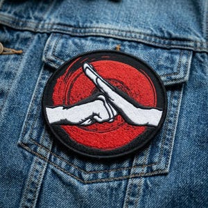 May include: A circular embroidered patch featuring a red background with a black circular border. The design depicts two white hands in a fist bump gesture, with one hand positioned horizontally and the other angled diagonally across the center.