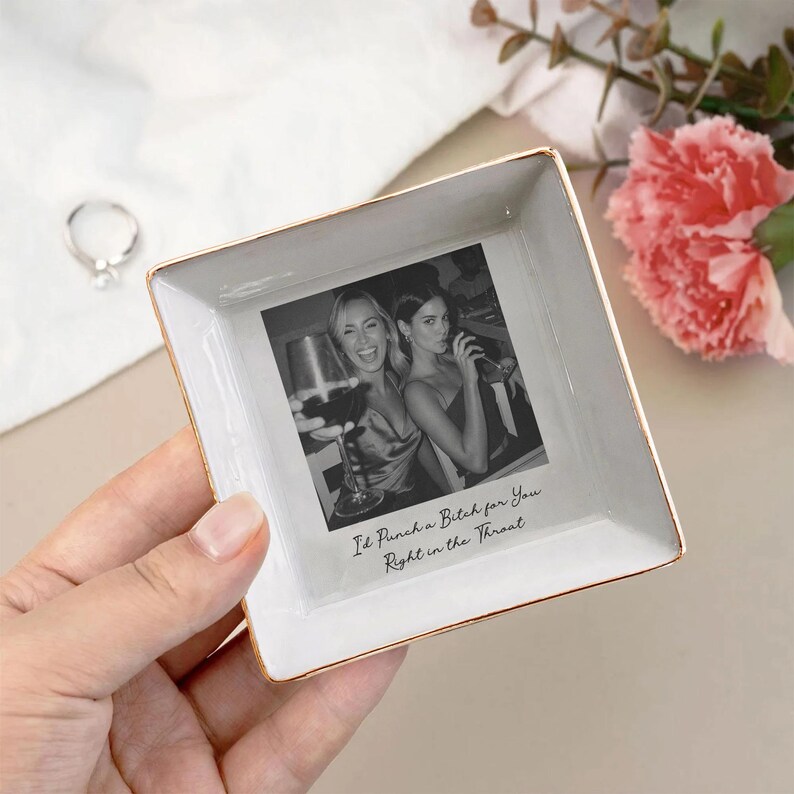 May include: Square white ceramic trinket dish with gold trim. The dish features a black and white photo of two women and the text "I'd Punch a Bitch for You Right in the Throat."