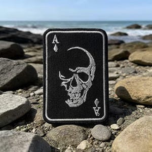 May include: Black rectangular patch featuring a skull design, resembling the Ace of Spades playing card. The skull is white, with details embroidered on a black background. The patch has a white border and the Ace symbol in the corners.