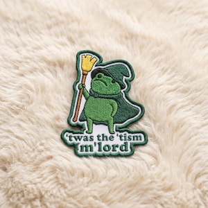 May include: An embroidered patch featuring a green frog wearing a dark green wizard hat and cape, holding a yellow broom. Below the frog, white text on a dark green background reads: 'twas the 'tism m'lord. The patch has a dark green border.