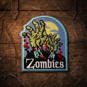 May include: Embroidered patch featuring zombie hands reaching up from a dark landscape, with a full moon in the background. The word "Zombies" is stitched in white. The patch has a blue border and is set against a wooden background.