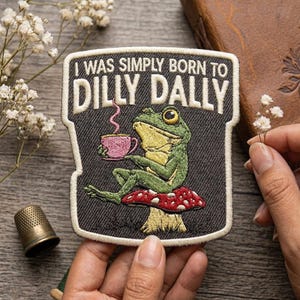 May include: Embroidered patch featuring a green frog sitting on a red and white mushroom, holding a pink teacup. The patch has the text "I WAS SIMPLY BORN TO DILLY DALLY" in white lettering on a dark gray background.