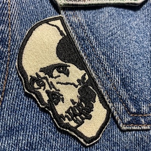 May include: Embroidered patch featuring a half-skull design in black and off-white. The patch is sewn onto a blue denim fabric, showcasing the detailed stitching and contrasting colors. The skull's features are clearly defined.