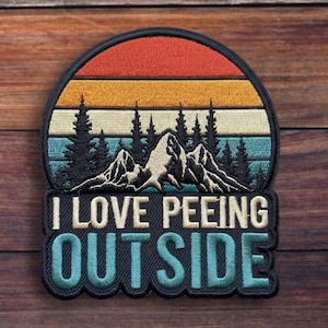 May include: Embroidered patch with a mountain and forest scene under a sunset. The patch reads "I LOVE PEEING OUTSIDE" in white and teal lettering. The patch is on a wooden background.