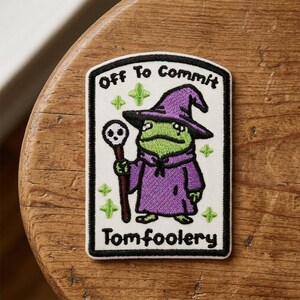May include: A rectangular embroidered patch with a white background and black border. It features a green frog wearing a purple wizard hat and robe, holding a staff with a skull topper. The text reads Off To Commit Tomfoolery with green stars.