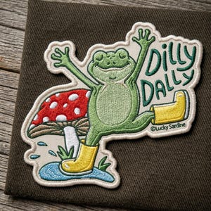 May include: Embroidered patch featuring a green frog wearing yellow boots, with arms raised in a joyful pose. A red and white mushroom and the words "Dilly Dally" are also included in the design. The patch is on a brown background.