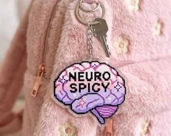May include: A keychain with a pixelated brain design in shades of purple and pink, with the words "NEURO SPICY" in white. The keychain is attached to a silver key and chain, hanging against a pink fuzzy backpack.