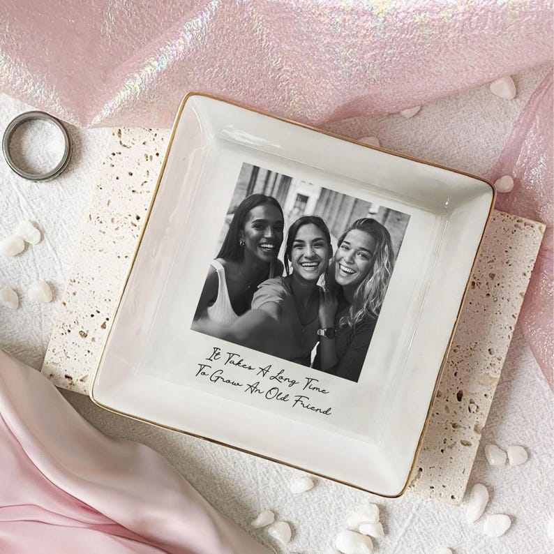 May include: Square white ceramic trinket tray with a gold rim, featuring a black and white photo of three smiling people. The text "It Takes A Long Time To Grow An Old Friend" is printed below the photo. A silver ring is visible.