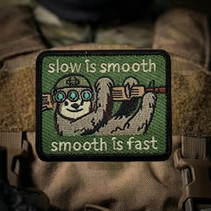 May include: A green and brown embroidered patch featuring a sloth wearing a helmet and holding a rifle. The text "slow is smooth, smooth is fast" is displayed in white. The patch is rectangular.