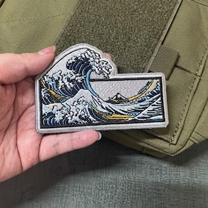 May include: Embroidered patch featuring the iconic Japanese artwork "The Great Wave off Kanagawa." The design showcases a large, curling wave with a boat and Mount Fuji in the background. The patch has a rectangular shape with a gray border and is attached to a green fabric.