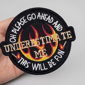 May include: Black embroidered patch with the text "OH PLEASE GO AHEAD AND UNDERESTIMATE ME THIS WILL BE FUN". The patch features a flame design in red and yellow, with white and gold lettering.