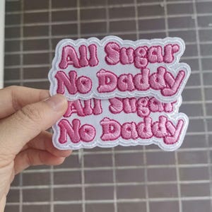 May include: Embroidered patch with the text "All Sugar No Daddy" in pink lettering on a white background. The patch has a white border and is held up by a hand.