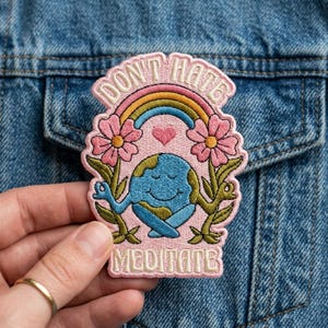 May include: Embroidered patch with the words "DON'T HATE MEDITATE" above a smiling Earth meditating under a rainbow. Pink flowers and green leaves frame the Earth. The patch is on a denim jacket.