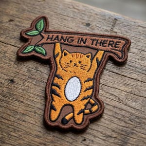 May include: An embroidered patch featuring an orange tabby cat with black stripes and a white belly, hanging from a brown tree branch. The branch includes green leaves and a banner with the text HANG IN THERE in black, capitalized letters.