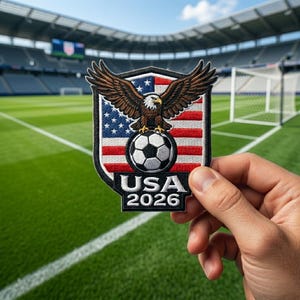 May include: A patch featuring a bald eagle perched atop a soccer ball, set against a backdrop of the American flag. The patch reads "USA 2026" in white text on a black background. The patch is held in front of a soccer field.