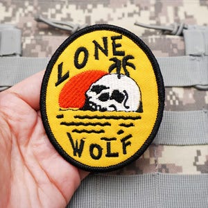 May include: A circular embroidered patch with a yellow background and a black border. The design features a white skull, a red sun, and a black palm tree above wavy lines. The text LONE WOLF is stitched in black lettering around the central graphic.