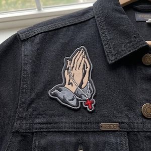 May include: A black denim jacket featuring an embroidered patch of two hands pressed together in prayer. The hands are light tan with grey sleeves, holding a white beaded rosary that ends in a small, bright red cross. The jacket has metal buttons.