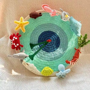 Op de afbeelding: Een gehaakte cirkelvormige wandhanger met een zeethema. Het midden is blauw, omgeven door concentrische ringen van groen en crème. Gehaakte zeedieren zijn onder meer een zeester, krab, octopus en garnaal.