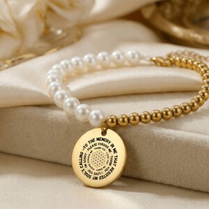May include: A gold-toned bracelet featuring a combination of white faux pearls and small, round gold beads. A circular pendant with engraved text hangs from the bracelet. The pendant has a floral design in the center and the text reads "Calling to the memory in me".