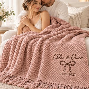 Könnte beinhalten: Eine altrosa, strukturiert gestrickte Wohndecke mit Fransen, die dunkelbraun bestickt ist mit dem Text Chloe & Owen, einer dekorativen Schleifengrafik und dem Datum 20.01.2027 in der unteren rechten Ecke des Stoffes.