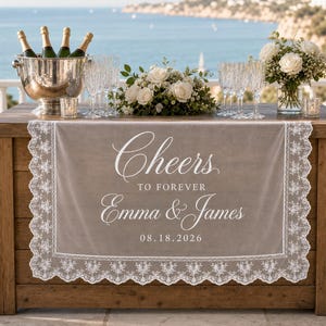May include: A beige table runner with white lace trim featuring the printed text Cheers TO FOREVER Emma & James 08.18.2026. The runner is displayed on a wooden table with a metal ice bucket, four wine bottles, clear glasses, and white flowers.