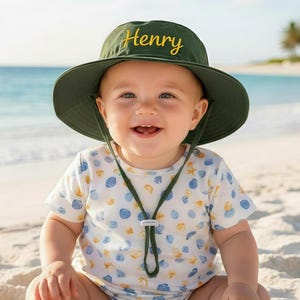 Puede incluir: Un bebé sonriente con un sombrero verde con la inscripción "Henry" en amarillo y un body blanco con un estampado de conchas marinas. El bebé está sentado en una playa de arena con el océano al fondo.