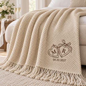 May include: A cream-colored textured knit throw blanket with fringe edges. The blanket features a dark brown embroidered design of two connected heart-shaped lockets with the letters M and R inside, followed by the date 05.22.2027 below.