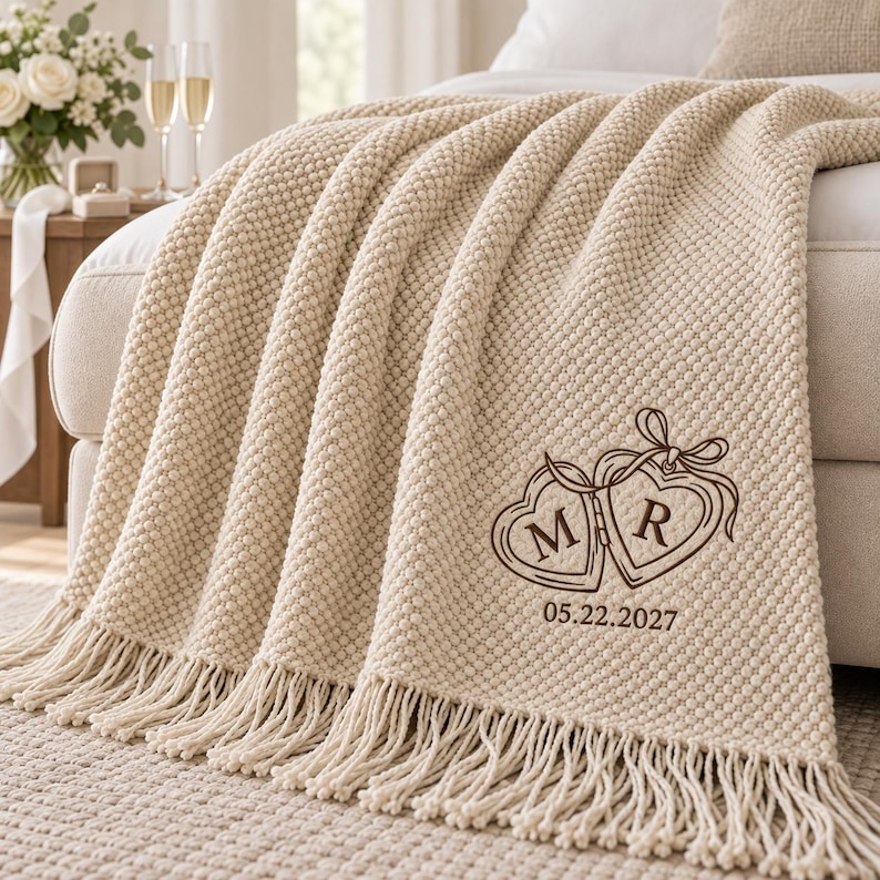 May include: A cream-colored textured knit throw blanket with fringe edges. The blanket features a dark brown embroidered design of two connected heart-shaped lockets with the letters M and R inside, followed by the date 05.22.2027 below.