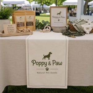 Op de afbeelding: Een beige tafelloper met de donkergroene tekst Poppy & Paw Natural Pet Goods, een hondensilhouet en een pootafdruk. De display bevat bruine zakjes met dierensnoepjes, een houten bordje, een ingelijst logo, een touwspeeltje en een groene hondenriem.
