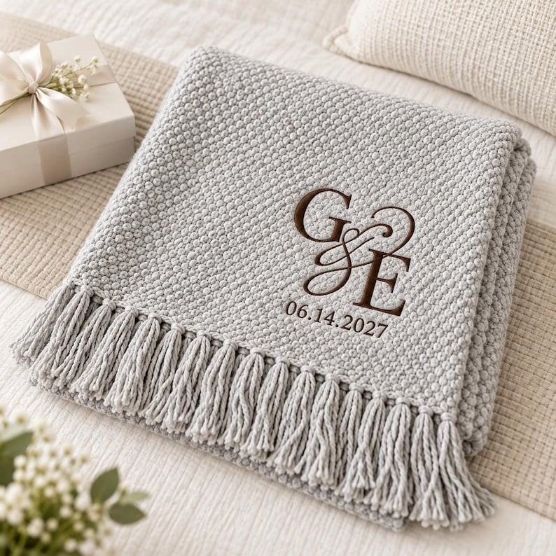 May include: A light gray knitted blanket with a textured popcorn stitch pattern and fringe trim along the edge. The blanket features dark brown embroidery of the letters G and E connected by an ampersand, with the date 06.14.2027 below it.