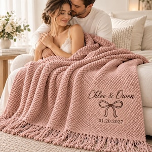 May include: A dusty rose textured knit throw blanket with fringe edges, featuring dark brown embroidered text that reads Chloe & Owen, a decorative bow graphic, and the date 01.20.2027 centered on the lower right corner of the fabric.