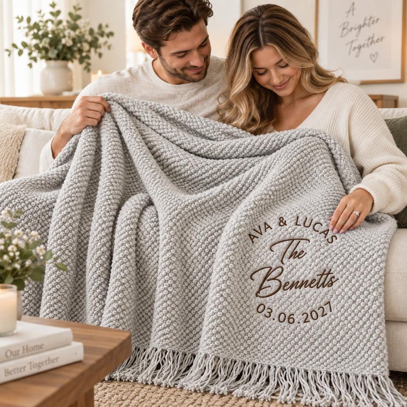 May include: A light gray textured throw blanket with fringe trim, featuring dark brown embroidered text that reads AVA & LUCAS, The Bennetts, and the date 03.06.2027 in a circular arrangement on the lower right corner of the fabric.