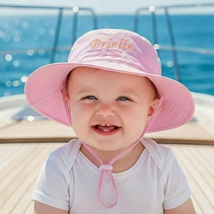 May include: A pink sun hat with the name "Brielle" embroidered on the front. The hat has a wide brim and a chin strap. The background shows a blue ocean and a wooden deck.