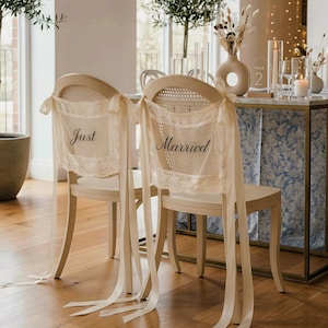 May include: Two cream-colored chairs with woven backs are adorned with lace and ribbon. The lace on the backs reads "Just Married" in black script. The chairs are part of a wedding decor setup.