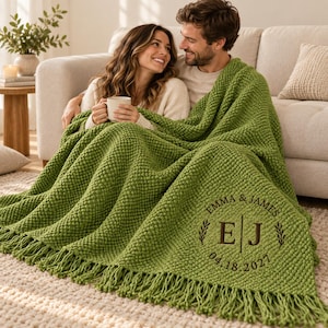 May include: A bright green chunky knit throw blanket with fringe trim. The blanket features a circular embroidered monogram with the text EMMA & JAMES, the initials E and J separated by a vertical line, and the date 04.18.2021 in dark brown thread.
