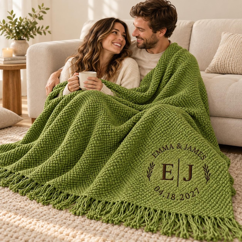 May include: A bright green chunky knit throw blanket with fringe trim. The blanket features a circular embroidered monogram with the text EMMA & JAMES, the initials E and J separated by a vertical line, and the date 04.18.2021 in dark brown thread.