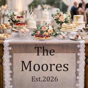 Peut inclure: Une table de desserts de mariage avec des gâteaux, des pâtisseries et des compositions florales. La table est recouverte d'un tissu transparent avec le texte "The Moores Est. 2026" et une bordure en dentelle blanche. Des desserts sont présentés sur des présentoirs en verre.