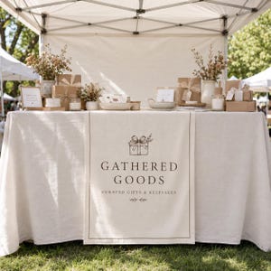 Puede incluir: Un camino de mesa color crema con el texto GATHERED GOODS CURATED GIFTS & KEEPSAKES y una ilustración de una caja de regalo. El camino se exhibe sobre una mesa con un mantel a juego, rodeado de cajas de regalo, jarrones y velas.