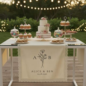 Könnte beinhalten: Ein Hochzeitsempfangstisch mit einem dreistöckigen Kuchen, der mit rosa Rosen verziert ist. Der Tisch zeigt auch Cupcakes, Kekse und Macarons. Ein cremefarbener Tischläufer mit "Alice & Ben" und dem Datum "05.18.2026" ist zu sehen.