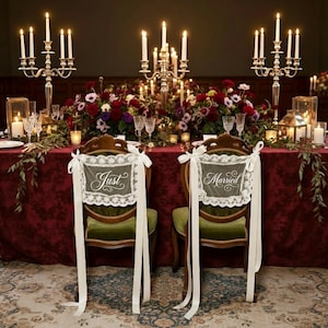 Puede incluir: Una romántica mesa de recepción de bodas. Dos sillas con letreros de "Just Married" y "Married" están frente a una mesa con un mantel burdeos, arreglos florales y candelabros. Las sillas son verdes con encaje blanco y cintas.