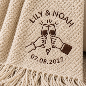 May include: A cream-colored textured throw blanket with fringe, featuring dark brown embroidery of two hands clinking champagne glasses, the text LILY & NOAH, a small heart, and the date 07.08.2027 centered on the fabric surface.