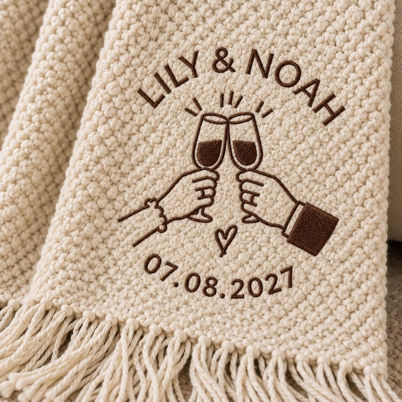 May include: A cream-colored textured throw blanket with fringe, featuring dark brown embroidery of two hands clinking champagne glasses, the text LILY & NOAH, a small heart, and the date 07.08.2027 centered on the fabric surface.