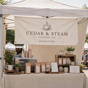 May include: A white canopy tent features a banner with the text CEDAR & STEAM COFFEE CO. Small Batch Coffee and a coffee cup icon. The booth displays a coffee machine, stacks of paper cups, brown coffee bags, a menu sign, and potted plants.