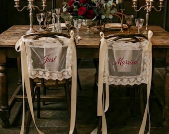 Op de afbeelding: Bruilofttafeldecoratie met twee stoelen versierd met doorschijnende witte stof en kant. De woorden "Just" en "Married" staan op de stof. De tafel is gedekt met kaarsen, bloemen en glaswerk.