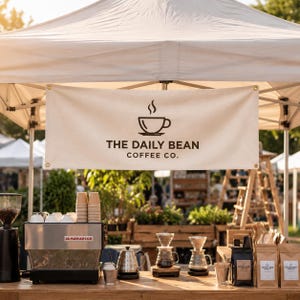 May include: A cream-coloured fabric banner featuring a dark brown logo of a coffee cup with steam and the text THE DAILY BEAN COFFEE CO. hanging under a white canopy, positioned above a professional espresso machine and coffee brewing equipment.