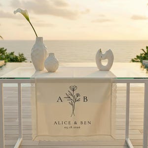 Puede incluir: Un camino de mesa de boda color crema con un diseño floral, iniciales y nombres. El camino de mesa está decorado con encaje y se muestra sobre una mesa de cristal con jarrones blancos y un lirio de agua. La fecha 05.18.2026 está impresa.