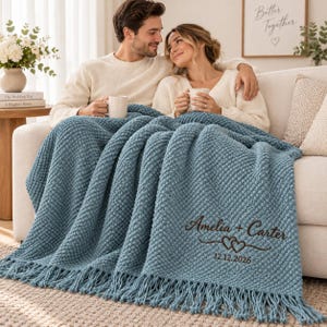 May include: A dusty blue textured throw blanket with fringe edges, featuring dark brown embroidered text that reads Amelia + Carter, a double heart graphic, and the date 12.12.2026. The blanket has a raised, bumpy knit pattern throughout its surface.